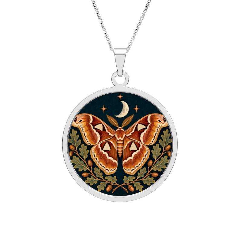 May include: A silver-toned circular pendant necklace featuring a detailed illustration of a large orange and brown moth centered against a dark background. The design includes a crescent moon, small stars, and green oak leaves with brown acorns.