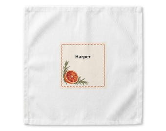 May include: A square white cloth featuring a centered cream-colored square label with a scalloped red border. The label displays the name Harper in black text, accompanied by a graphic of a sliced orange and green rosemary sprigs.