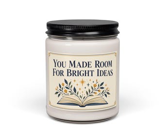 Puede incluir: Una vela blanca en un frasco de vidrio transparente con tapa negra. La etiqueta presenta el texto "YOU MADE ROOM FOR BRIGHT IDEAS" sobre una ilustración de un libro abierto con ramas florales y estrellas pequeñas.