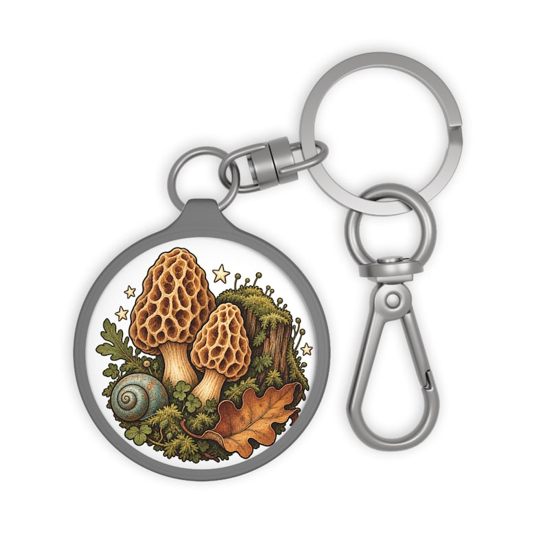 May include: A circular keychain featuring a silver-toned metal ring and clasp. The central disc displays a detailed illustration of two tan morel mushrooms, a small blue snail, a brown oak leaf, and green moss, accented by several small yellow stars.