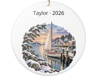 May include: A round white ceramic ornament featuring the text Taylor 2026. The design depicts a sailboat docked at a wooden pier during a snowy winter sunset, framed by snow-covered pine branches and rocks, with a distant lighthouse.