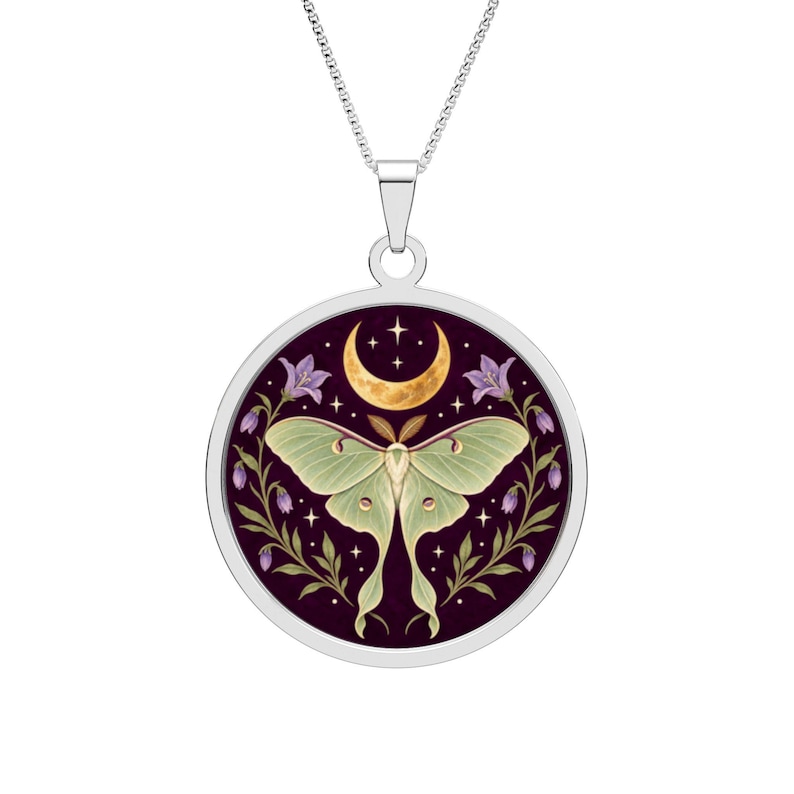 May include: A silver-toned circular pendant necklace featuring a central illustration of a pale green moth with long wings. The moth is framed by purple bell-shaped flowers against a dark purple background, topped with a golden crescent moon.