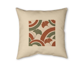 Op de afbeelding: Een vierkant sierkussen in een lichtbeige kleur met een gecentreerde grafische print. Het ontwerp bestaat uit herhalende halve cirkelbogen in terracotta oranje en saliegroen, afgewisseld met gestileerde illustraties van ginkgobladeren.