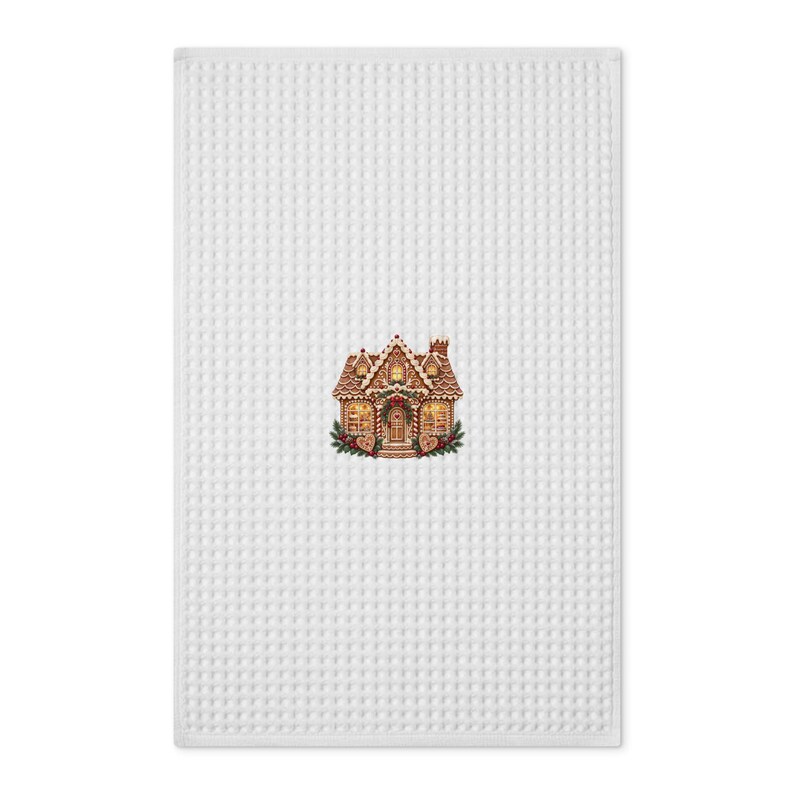 May include: A white waffle-weave kitchen towel featuring a centered graphic of a gingerbread house. The house is decorated with brown icing, red and white candy cane accents, and green foliage, with warm yellow light glowing from the windows.