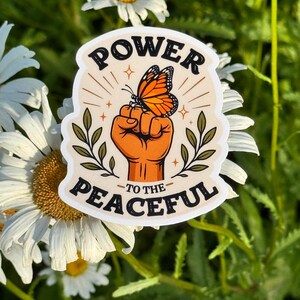 May include: A die-cut sticker featuring a raised fist in an orange tone with a monarch butterfly perched on top. The design includes green leafy branches on both sides and the black text "POWER TO THE PEACEFUL" arched above and below the central image.