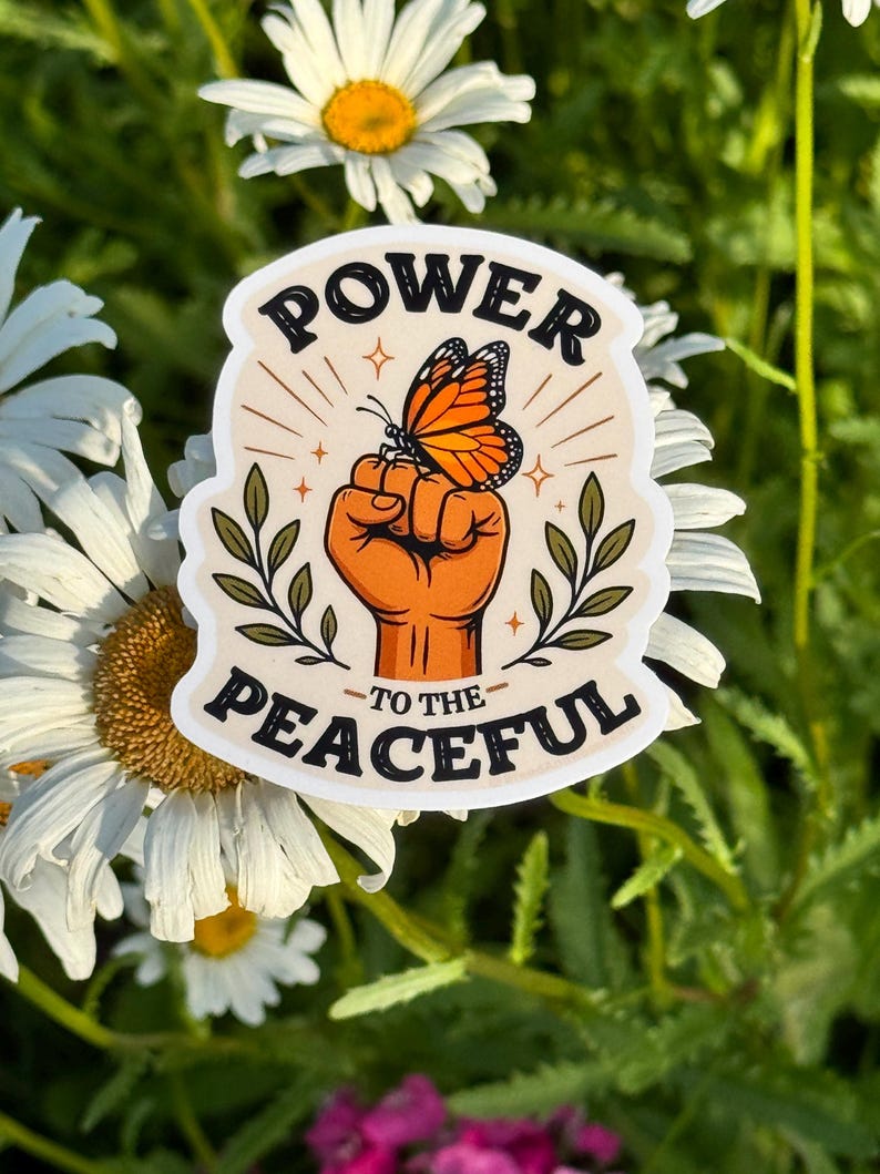 May include: A die-cut sticker featuring a raised fist in an orange tone with a monarch butterfly perched on top. The design includes green leafy branches on both sides and the black text "POWER TO THE PEACEFUL" arched above and below the central image.