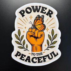 May include: A die-cut sticker featuring a raised fist in orange with a monarch butterfly perched on the knuckles. The design includes green leafy branches on both sides, gold star accents, and the black text POWER TO THE PEACEFUL on a cream background.