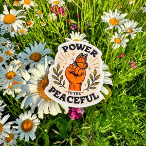 May include: A die-cut sticker featuring an orange fist raised upward with a monarch butterfly perched on the knuckles. The design includes green leafy branches on both sides and the black text "POWER TO THE PEACEFUL" arched around the illustration.