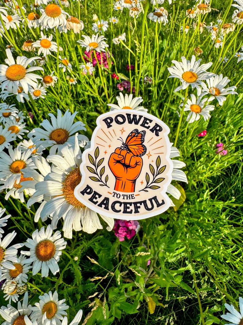 May include: A die-cut sticker featuring an orange fist raised upward with a monarch butterfly perched on the knuckles. The design includes green leafy branches on both sides and the black text "POWER TO THE PEACEFUL" arched around the illustration.