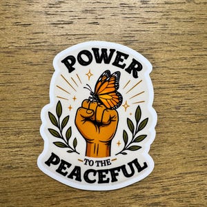 May include: A die-cut sticker featuring a raised fist in a warm orange tone with a monarch butterfly perched on the knuckles. The text POWER TO THE PEACEFUL is printed in black, with green leafy branches on both sides and small gold star accents.