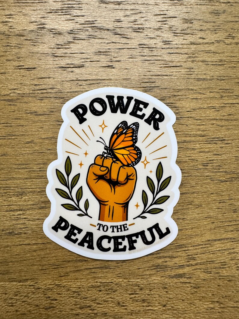 May include: A die-cut sticker featuring a raised fist in a warm orange tone with a monarch butterfly perched on the knuckles. The text POWER TO THE PEACEFUL is printed in black, with green leafy branches on both sides and small gold star accents.