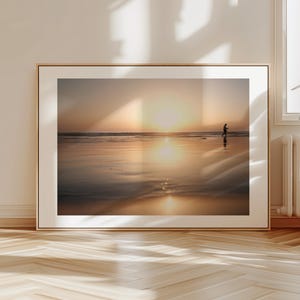 Puede incluir: Fotografía enmarcada de una escena de playa al atardecer. La imagen presenta a una persona caminando sobre la arena mojada, con el sol bajo en el horizonte. El cielo y el agua reflejan los cálidos colores del atardecer.