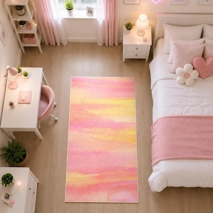 Pode incluir: Um tapete retangular com um padrão de gradiente suave em estilo aguarela em tons de rosa, pêssego e amarelo. O tapete está posicionado sobre um chão de madeira clara no centro de um quarto, exibindo uma mistura horizontal de cores pastel.