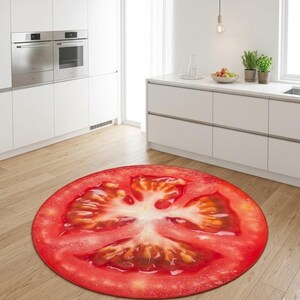 May include: A circular rug depicting a realistic tomato slice, with red and yellow colours. The rug is on a wooden floor in a modern kitchen, with white cabinets and stainless steel appliances visible in the background.