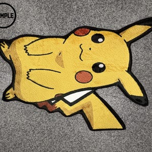 May include: A yellow Pikachu-shaped rug with black outlines and brown accents. The rug features the text "SAMPLE" in a black circle. The rug is on a grey surface.