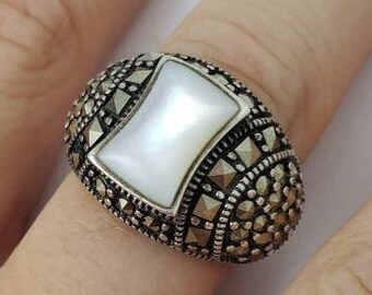 May include: A silver ring with a white mother-of-pearl stone set in a square shape surrounded by marcasite stones.