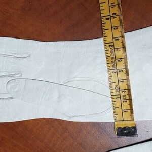 K&ouml;nnte beinhalten: Ein Paar wei&szlig;e Lederhandschuhe mit einem kleinen Riss am B&uuml;ndchen. Die Handschuhe werden mit einem Lineal gemessen, das bis zu 28 cm misst.