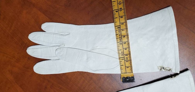 K&ouml;nnte beinhalten: Ein Paar wei&szlig;e Lederhandschuhe mit einem kleinen Riss am B&uuml;ndchen. Die Handschuhe werden mit einem Lineal gemessen, das bis zu 28 cm misst.