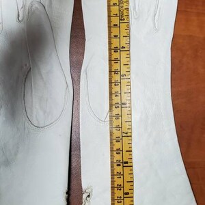 K&ouml;nnte beinhalten: Zwei Paar wei&szlig;e Lederhandschuhe mit einer Naht in der Mitte jedes Fingers. Die Handschuhe liegen flach auf einer Holzoberfl&auml;che mit einem Lineal, das 23 cm misst.