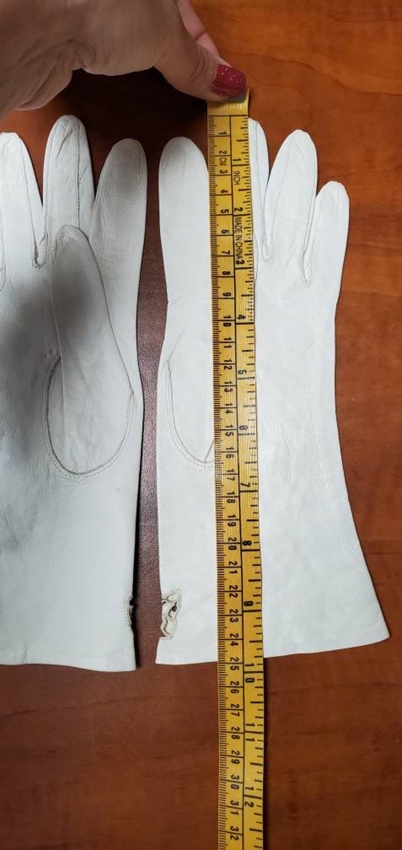 K&ouml;nnte beinhalten: Zwei Paar wei&szlig;e Lederhandschuhe mit einer Naht in der Mitte jedes Fingers. Die Handschuhe liegen flach auf einer Holzoberfl&auml;che mit einem Lineal, das 23 cm misst.