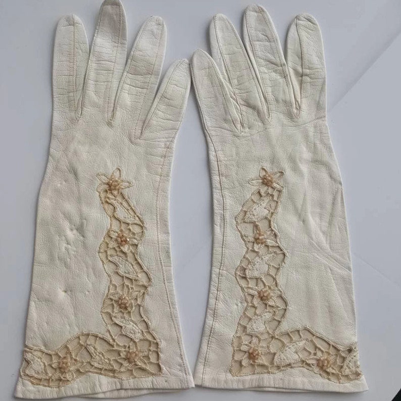 K&ouml;nnte beinhalten: Ein Paar wei&szlig;e Lederhandschuhe mit zarten Spitzen- und Perlenverzierungen. Die Handschuhe sind lang und reichen &uuml;ber das Handgelenk.