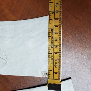 K&ouml;nnte beinhalten: Ein wei&szlig;er Lederhandschuh mit einer Nahtdetail auf der R&uuml;ckseite der Hand. Der Handschuh wird mit einem Lineal gemessen, das "2 cm" bis "11" Zoll anzeigt.