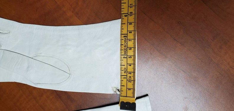 K&ouml;nnte beinhalten: Ein wei&szlig;er Lederhandschuh mit einer Nahtdetail auf der R&uuml;ckseite der Hand. Der Handschuh wird mit einem Lineal gemessen, das "2 cm" bis "11" Zoll anzeigt.