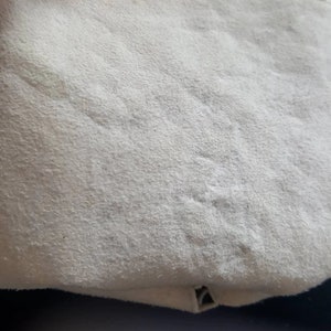 K&ouml;nnte beinhalten: Nahaufnahme einer wei&szlig;en Lederoberfl&auml;che mit subtiler Textur. Die Oberfl&auml;che scheint Teil eines Kleidungsst&uuml;cks oder Accessoires zu sein.