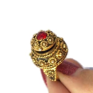 May include: A gold ring featuring a red jewel set into the top. The ring is intricately detailed with a floral design and has a hinged lid that opens to reveal a small compartment.