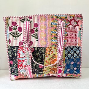 May include: A patchwork tote bag with a variety of floral and geometric patterns in pink, red, black, and blue. The bag has a quilted texture and a pink and white striped trim. The handles are made of the same fabric as the trim.