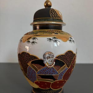 Op de afbeelding: Een decoratieve keramische pot met een deksel. De pot heeft een witte basis met een geschilderde scène van een persoon in traditionele kleding. Het deksel is zwart met gouden accenten en de pot heeft een gouden rand.