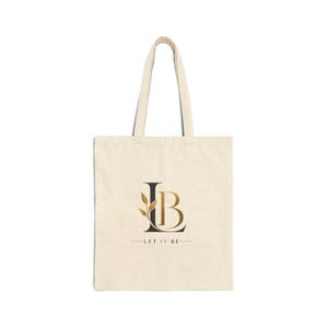 May include: A beige canvas tote bag featuring a black and gold graphic design on the front. The design displays the letters LB with a gold leaf branch accent on the L, and the text LET IT BE centered underneath with horizontal lines on both sides.