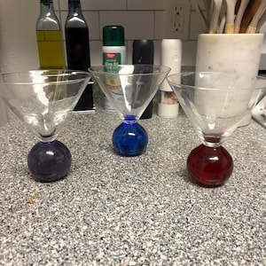 May include: Three clear martini glasses with colored bases: purple, blue, and red. The glasses are on a speckled countertop, with kitchen items in the background. The glasses have a classic martini shape.