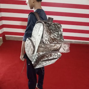 Puede incluir: Una mochila hecha de piel de vaca con un patr&oacute;n moteado de pelo blanco y marr&oacute;n oscuro. La bolsa incluye un compartimento principal grande con una solapa superior, un bolsillo frontal m&aacute;s peque&ntilde;o y correas y adornos de cuero marr&oacute;n con cremalleras.