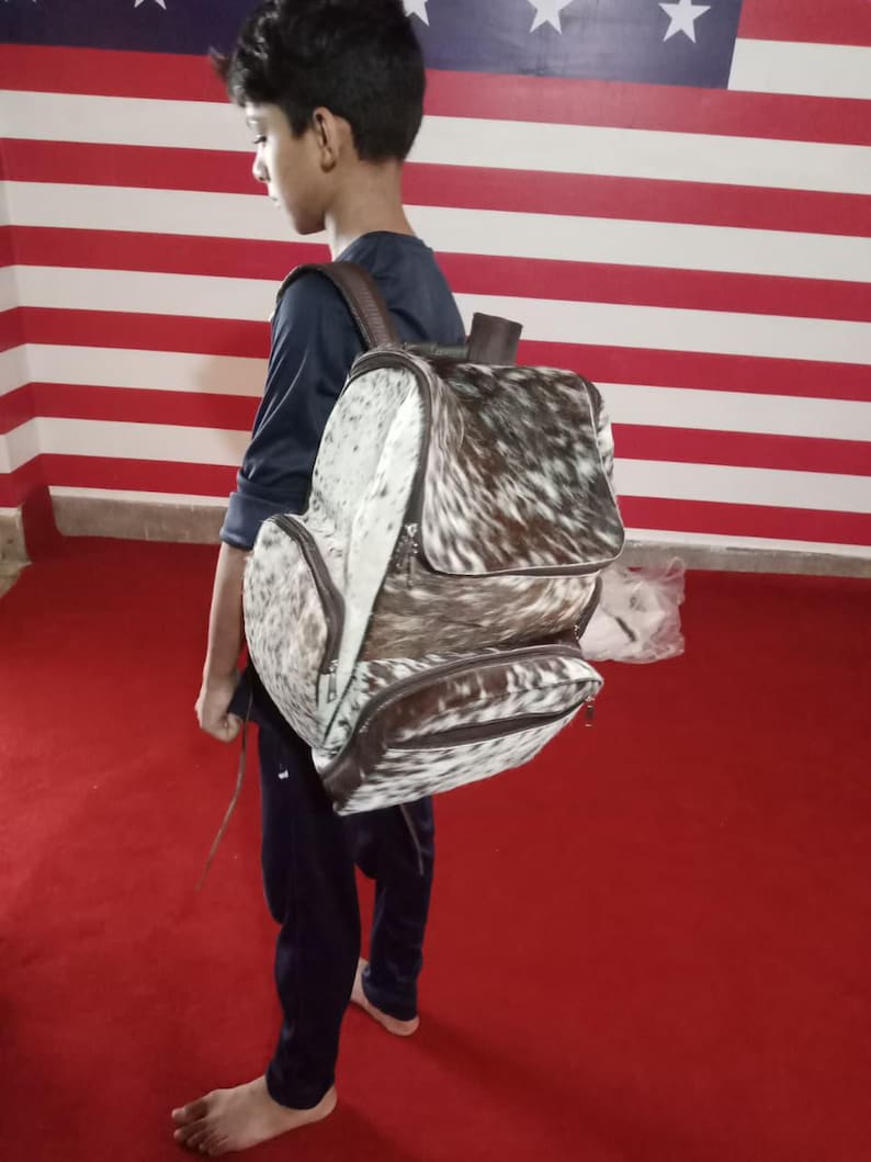 Puede incluir: Una mochila hecha de piel de vaca con un patr&oacute;n moteado de pelo blanco y marr&oacute;n oscuro. La bolsa incluye un compartimento principal grande con una solapa superior, un bolsillo frontal m&aacute;s peque&ntilde;o y correas y adornos de cuero marr&oacute;n con cremalleras.