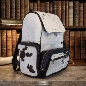 Puede incluir: Una mochila confeccionada en piel de vaca blanca y negra con un compartimento principal grande con solapa, un bolsillo frontal con cremallera y ribetes de cuero negro. El bolso muestra un patr&oacute;n de manchas natural y correas negras.
