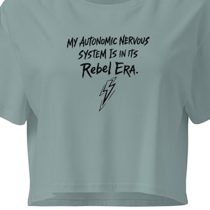 Puede incluir: Una camiseta corta de color verde azulado claro con el texto negro "MY AUTONOMIC NERVOUS SYSTEM IS IN ITS Rebel ERA." y un rayo. La camiseta tiene mangas cortas y cuello redondo.