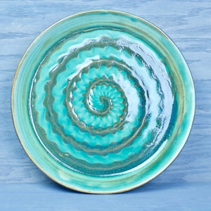 May include: A handmade ceramic plate with a turquoise spiral design. The plate has a glossy finish and a golden rim. The spiral pattern is a darker shade of turquoise, creating a contrast against the lighter background. The plate is a unique piece of art.