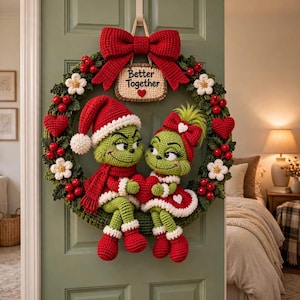 May include: A crochet wreath featuring two green characters with red and white clothing, sitting together and holding a red heart. The wreath is decorated with red berries, white flowers, and red hearts. A sign at the top reads Better Together.
