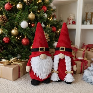May include: Two handmade crochet gnomes featuring tall red pointed hats with black belts and gold buckles. One gnome has a long white yarn beard, while the other has two long white braided pigtails. Both figures have beige noses and black feet.