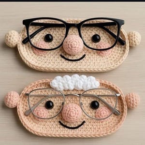 Peut inclure: Deux porte-lunettes ovales en crochet beige, chacun doté d'yeux en perles noires, d'un nez rose rond et d'un sourire brodé. Le support supérieur présente des lunettes à monture noire, tandis que l'inférieur porte des lunettes argentées et des cheveux blancs.