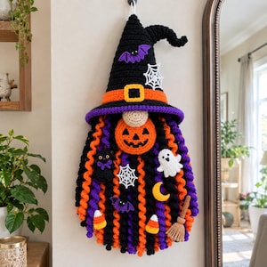 May include: A crocheted wall hanging featuring a gnome with a tall black witch hat, an orange pumpkin face, and a long beard made of spiraled black, orange, and purple yarn. The beard is decorated with small crocheted ghosts, bats, spiders, webs, candy corn, a moon, and a broom.
