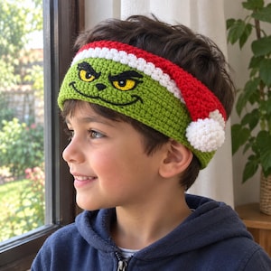 May include: A green crochet headband featuring a character face with yellow eyes and a black stitched smile. The top edge has a red and white trim, and a white pom-pom is attached to the side, resembling a festive holiday hat design.