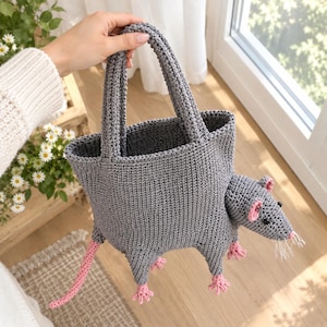 Puede incluir: Un bolso de mano tejido a ganchillo en color gris con forma de rata. El bolso presenta un cuerpo gris texturizado, dos asas superiores, una larga cola rosa, cuatro pequeñas patas rosas y una cabeza de rata con ojos negros, orejas rosas y bigotes.