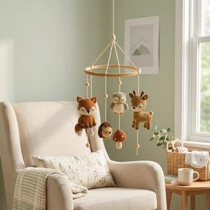 May include: A hanging nursery mobile featuring a circular wooden frame with five crocheted figures suspended by white strings: a brown fox, a grey owl, a brown hedgehog, a red mushroom, and a brown deer, all crafted with yarn.
