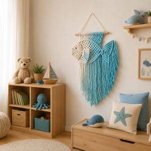 May include: A macrame wall hanging shaped like a fish, featuring a gradient of cream and light blue cotton cord. The fish design includes a knotted body, a fin, and long, flowing fringe strands suspended from a horizontal wooden dowel.