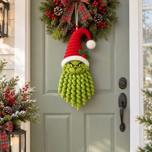 May include: A crochet wall hanging featuring a green character with a long curly beard, wearing a red and white hat with a white pom-pom and holly leaves. The character has yellow eyes and a wide grin, suspended from a festive evergreen wreath.