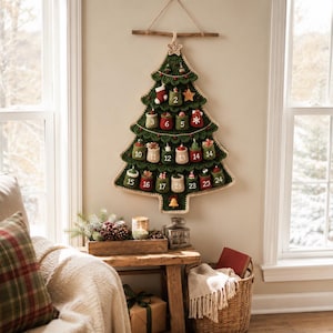 Könnte beinhalten: Ein gehäkelter Adventskalender in Form eines Weihnachtsbaums in Grüntönen, mit vierundzwanzig kleinen nummerierten Taschen und verschiedenen Miniaturornamenten wie einem Strumpf, einem Stern und einer Glocke, aufgehängt an einem Holzstab.