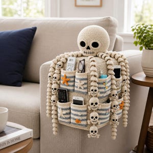 May include: A beige crochet organiser designed to drape over a sofa arm, featuring a skull head at the top and long tentacles adorned with small skulls. It includes multiple pockets with blue and white stripes, decorated with starfish and shells.