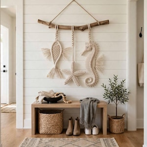 May include: A macrame wall hanging featuring three off-white knotted figures—a fish, a starfish, and a seahorse—suspended from a horizontal piece of driftwood by braided cords. The intricate fiber art displays detailed knot patterns.
